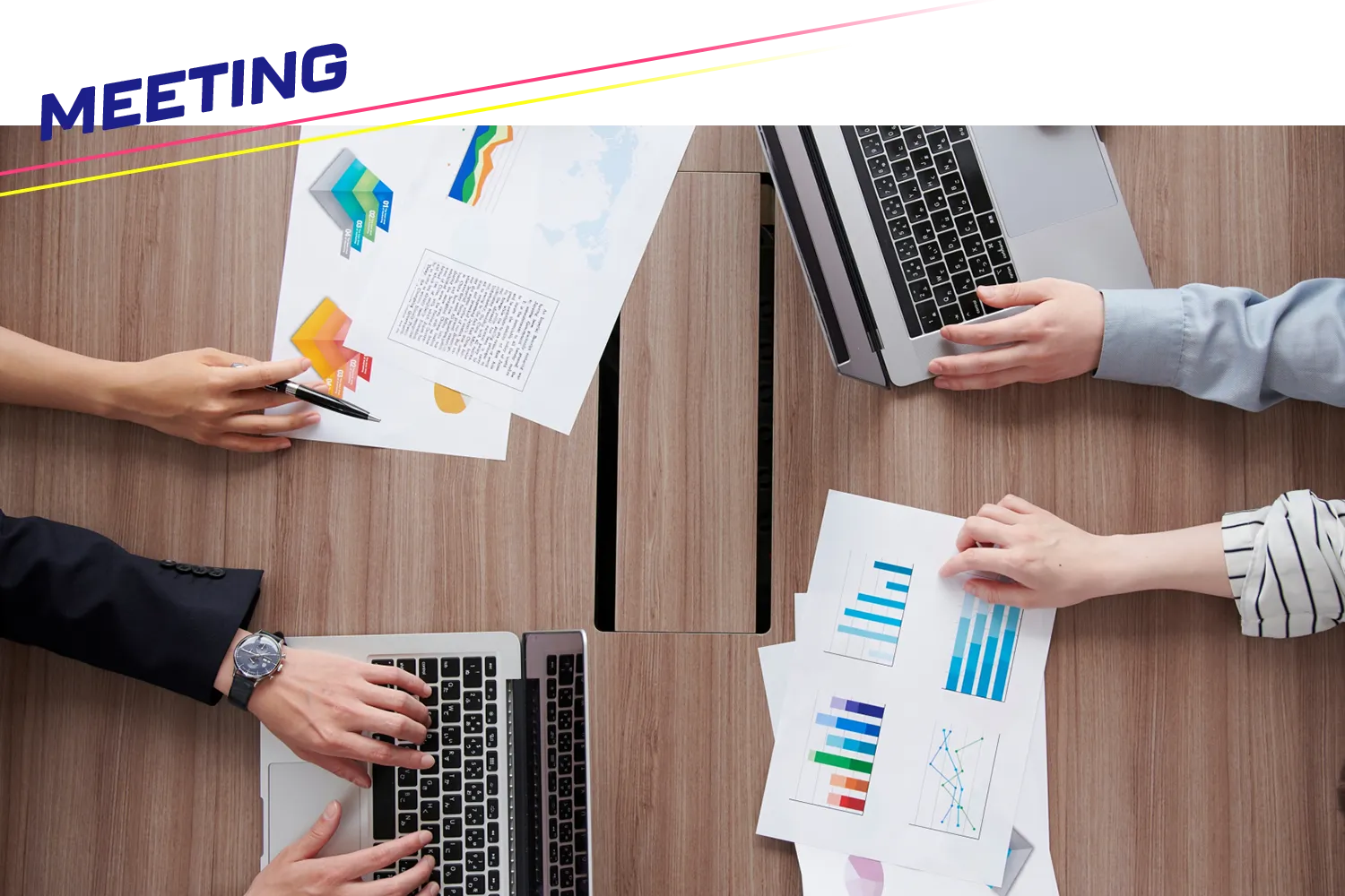 パソコンと資料と人の手が乗った会議テーブルを上から見た写真